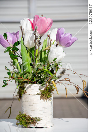 Beautiful tulips in purple, white, and pink are displayed in a rustic vase, surrounded by fresh greenery and moss, evoking spring 122976657