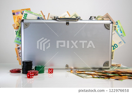 A silver briefcase filled with Euro and US dollar bills sits on a white table surrounded by casino chips and dice A silver briefcase filled with Euro and US dollar bills sits on a white table surrounded by casino chips and dice 122976658