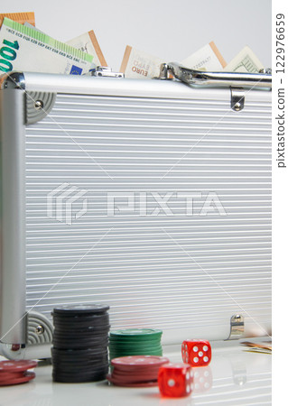 A silver briefcase with euro bills sticking out of the top. Poker chips and red dice are scattered on the white table in front of the case 122976659
