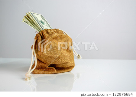 A small, brown burlap bag sits on a white surface. The bags drawstring is pulled tight, and a bundle of US dollar bills is peeking out from the top A small, brown burlap bag sits on a white surface. The bags drawstring is pulled tight, and a bundle of US dollar bills is peeking out from the top 122976694
