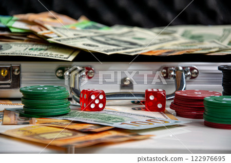 Close-up of red dice and stacks of poker chips on a white surface. A silver suitcase filled with money is visible in the background Close-up of red dice and stacks of poker chips on a white surface. A silver suitcase filled with money is visible in the background 122976695