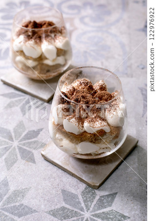 Classic Italian tiramisu dessert served in two glasses. Layers of coffee-soaked sponge and creamy mascarpone, dusted with cocoa powder. Placed on patterned gray-blue ceramic tile, spoon beside 122976922