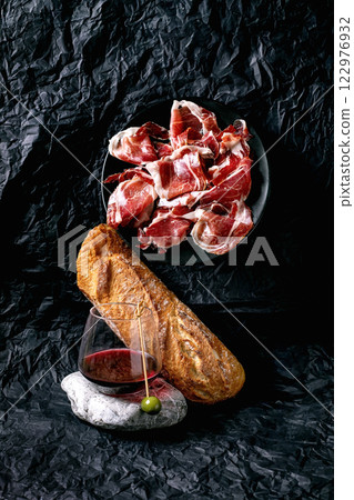 Still life with sliced Spanish ham Jamon Iberico, paleta on plate, glass of red wine with olive on stone, white bread loaf. Crumpled black paper background. Concept of traditional Spanish cuisine. 122976932