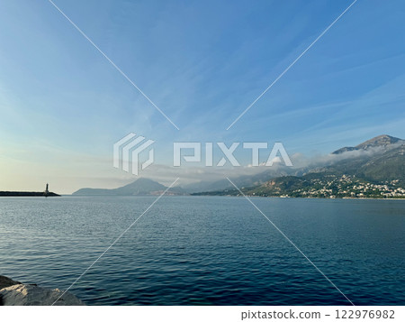 A panoramic view of Bar, Montenegro, in warm pre-sunset light with clear blue sky snd seascape, a distant horizon line, a lighthouse, and mountains in the background 122976982