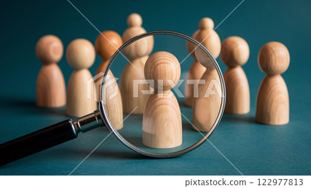 One wooden figurine among many others on a teal background is the subject of a magnifying glass. One wooden figurine among many others on a teal background is the subject of a magnifying glass. 122977813