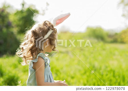 Easter egg hunt. Girl child Wearing Bunny Ears Running To Pick Up Egg In Garden. Easter tradition. Baby with basket full of colorful eggs. 122978078