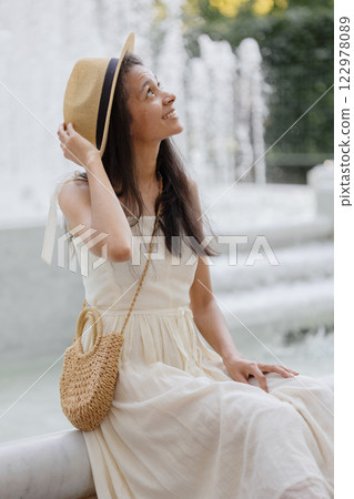young African American woman in white dress and straw hat sits by white marble fountain. young African American woman in white dress and straw hat sits by white marble fountain. 122978089
