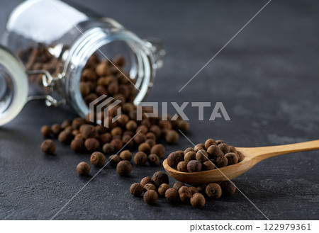 allspice or Jamaican pepper in a wooden scoop and in glass storage jar on a black kitchen table, selective focus. allspice or Jamaican pepper in a wooden scoop and in glass storage jar on a black kitchen table, selective focus. 122979361