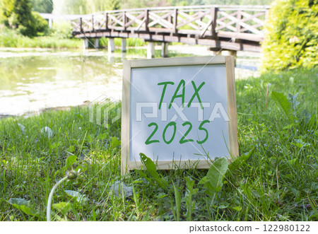 Tax 2025 symbol. White Blackboard with words Tax 2025. Beautiful meadow background with bridge. Business and Tax 2025 concept. Copy space. Tax 2025 symbol. White Blackboard with words Tax 2025. Beautiful meadow background with bridge. Business and Tax 2025 concept. Copy space. 122980122