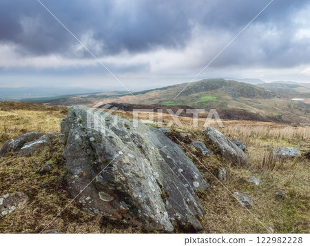 A bleak, hilly landscape presents itself. A grey sky and scattered rocks are visible. The distant hills appear under the dark clouds. A bleak, hilly landscape presents itself. A grey sky and scattered rocks are visible. The distant hills appear under the dark clouds. 122982228