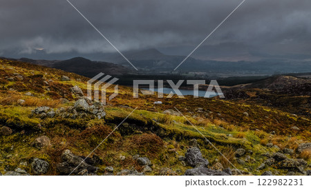 A vast landscape showcases a serene lake surrounded by majestic mountains, shrouded in dark clouds and mossy terrain, capturing a tranquil yet moody atmosphere in nature. 122982231