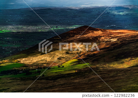 Majestic green hills illuminated by sunlight in a serene landscape during late afternoon. Majestic green hills illuminated by sunlight in a serene landscape during late afternoon. 122982236