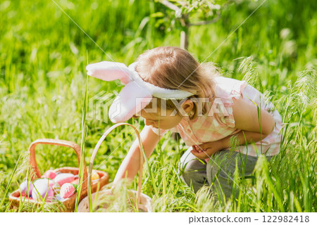 Easter egg hunt. Girl child Wearing Bunny Ears Running To Pick Up Egg In Garden. Easter tradition. Baby with basket full of colorful eggs. 122982418