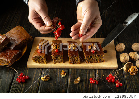 A pair of hands meticulously arranges vibrant red berries viburnum and walnuts atop rich cake slices. The rustic wooden table enhances the warm, inviting feel of this baking moment A pair of hands meticulously arranges vibrant red berries viburnum and walnuts atop rich cake slices. The rustic wooden table enhances the warm, inviting feel of this baking moment 122983397