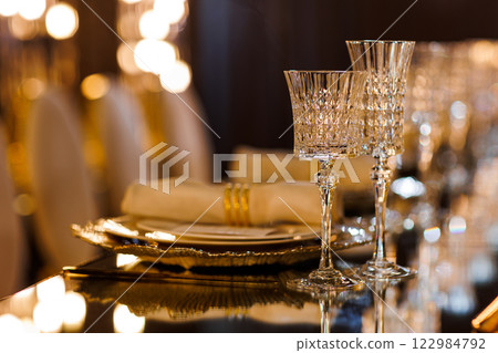 Luxury table setting with plates, napkin with ring, crystal wineglasses and golden cutlery on mirror table for romantic dinner in restaurant, bokeh. Christmas, New Year 2026 dinner. Wedding, birthday Luxury table setting with plates, napkin with ring, crystal wineglasses and golden cutlery on mirror table for romantic dinner in restaurant, bokeh. Christmas, New Year 2026 dinner. Wedding, birthday 122984792