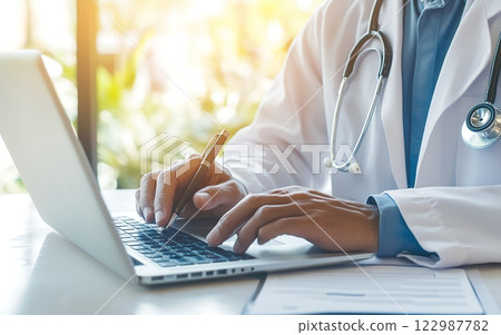 A professional moment for a doctor in a white coat working on a laptop at his desk A professional moment for a doctor in a white coat working on a laptop at his desk 122987782