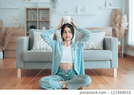 Young Asian woman holding piggy bank and sitting on the floor at home Young Asian woman holding piggy bank and sitting on the floor at home 122989018