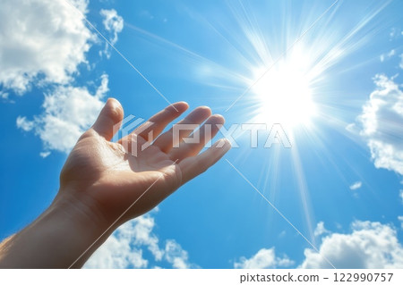 Hand Reaching Toward Bright Sunlight in Blue Sky with Rays and White Fluffy Clouds 122990757