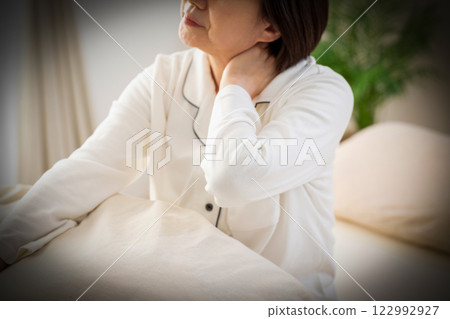 一名身穿白色睡衣的中年婦女早上在臥室裡感到頸部和肩膀僵硬 122992927
