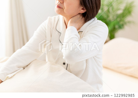 一名身穿白色睡衣的中年婦女早上在臥室裡感到頸部和肩膀僵硬 122992985