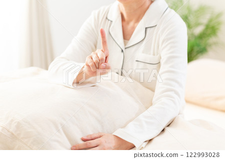 A middle-aged woman in white pajamas pointing up in her bedroom in the morning 122993028