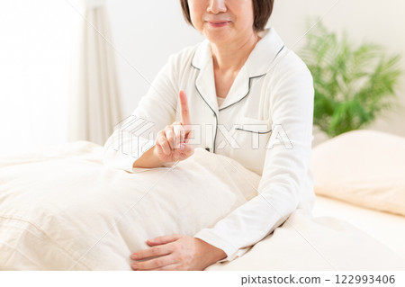 A middle-aged woman in white pajamas pointing up in her bedroom in the morning 122993406