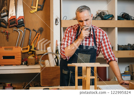 Senior carpenter in plaid shirt and overalls inspects a wooden stool in a workshop, surrounded by tools and materials. Workshop wall with tools visible in the background, showcasing craftsmanship Senior carpenter in plaid shirt and overalls inspects a wooden stool in a workshop, surrounded by tools and materials. Workshop wall with tools visible in the background, showcasing craftsmanship 122995824