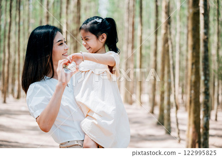 Asian mother carrying a daughter in arms in a peaceful forest, Happy smile mother and little child make heart sign with hands enjoy close tender moment together, Family day memory on holiday concept 122995825
