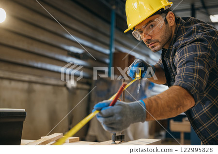 Carpenter wearing a yellow hard hat and safety glasses measuring wood with tape measure. Ideal for woodworking, furniture making, and cabinetry, showcasing precision 122995828
