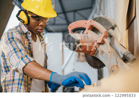 Construction worker in a yellow helmet using a chop saw to cut wood, with a vacuum cleaner collecting wood chips. woodworking, precision cutting, and construction sites, National Carpenters Day Construction worker in a yellow helmet using a chop saw to cut wood, with a vacuum cleaner collecting wood chips. woodworking, precision cutting, and construction sites, National Carpenters Day 122995833