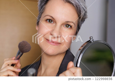 Mature happy beautiful woman during menopause with grey hair applies makeup to her face 122998656