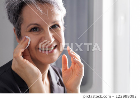Mature well-groomed beautiful woman during menopause with grey hair removes daytime makeup 122999092