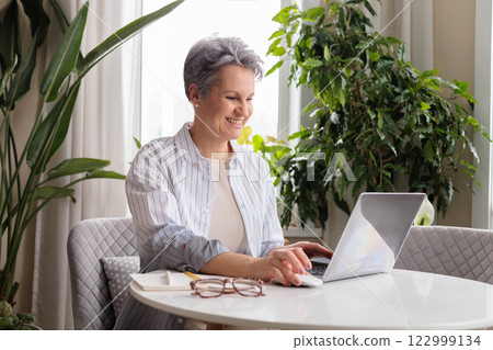 Smiling stylish mature middle aged woman sitting at table with laptop, portrait. Smiling stylish mature middle aged woman sitting at table with laptop, portrait. 122999134