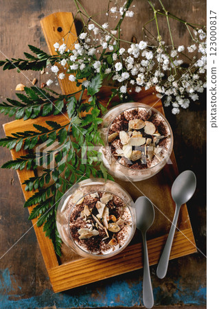Classic Italian tiramisu layered dessert with cocoa powder and almonds in two glasses. Served on tray with spoons on old wooden table. Decorated white babys breath flowers and green leaves. Top view 123000817