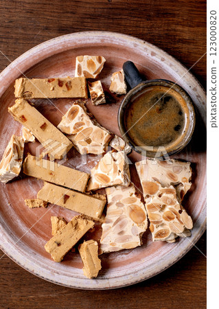 Traditional Spanish dessert classic soft orange turron and crunchy almond turron, cut into pieces on rustic clay plate. Espresso in black cup, wooden table, top view. 123000820