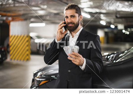 Young Caucasian male businessman in suit talking on phone holding cash in underground parking lot. Young Caucasian male businessman in suit talking on phone holding cash in underground parking lot. 123001474