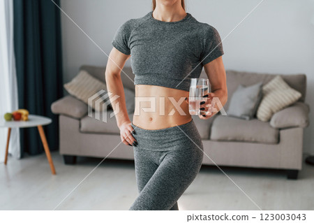 With glass of water. Young woman with slim body type and in yoga clothes is at home With glass of water. Young woman with slim body type and in yoga clothes is at home 123003043