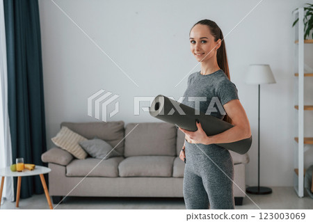Standing with mat in hands. Young woman with slim body type and in yoga clothes is at home Standing with mat in hands. Young woman with slim body type and in yoga clothes is at home 123003069
