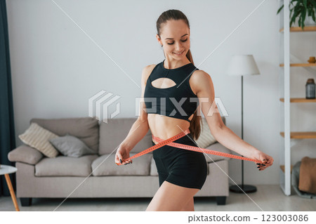 Measuring the waist by the tape. Young woman with slim body type and in yoga clothes is at home 123003086