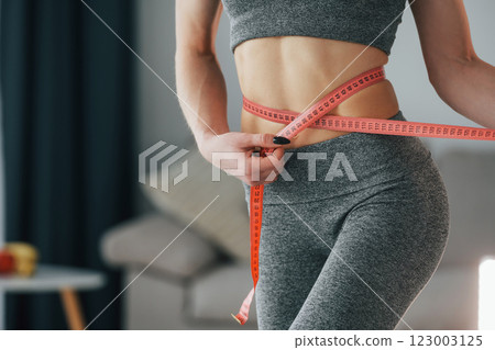 Pink colored measuring tape around the waist. Young woman with slim body type and in yoga clothes is at home 123003125
