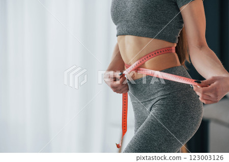 Pink colored measuring tape around the waist. Young woman with slim body type and in yoga clothes is at home 123003126
