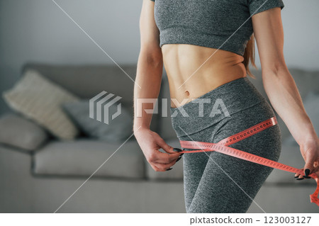 Pink colored measuring tape around the waist. Young woman with slim body type and in yoga clothes is at home 123003127