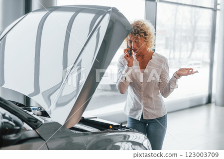 Car is damaged, hood is opened. Woman with curly blonde hair is in autosalon 123003709