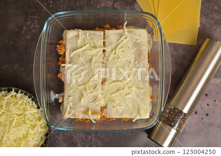 Top view. Homemade lasagna cooking preparation. Lasagne pasta sheets, Bolognese sauce in a glass form. Grated cheese in a black bowl, pepper mill. Black background 123004250