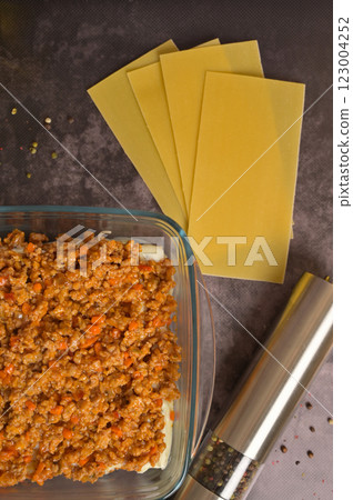 Top view. Bolognese sauce in a glass form. Lasagne pasta sheets, pepper mill. Black background. Homemade lasagna cooking preparation 123004252