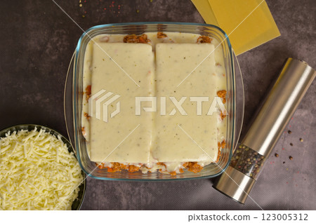 Top view. Homemade lasagna cooking preparation. Lasagne pasta sheets, Bolognese sauce in a glass form. Grated cheese in a black bowl, pepper mill. Black background 123005312