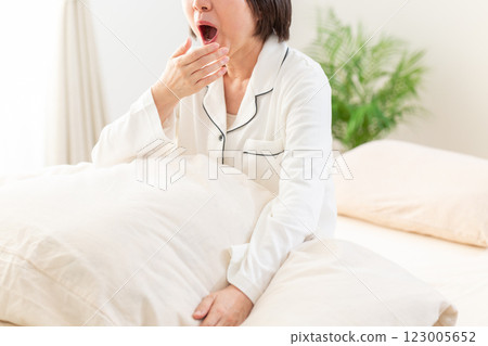 A middle-aged woman in white pajamas yawning in her bedroom in the morning 123005652