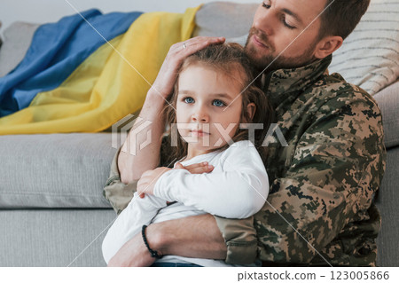 Ukrainian flag is on the sofa. Soldier in uniform is at home with his little daughter Ukrainian flag is on the sofa. Soldier in uniform is at home with his little daughter 123005866