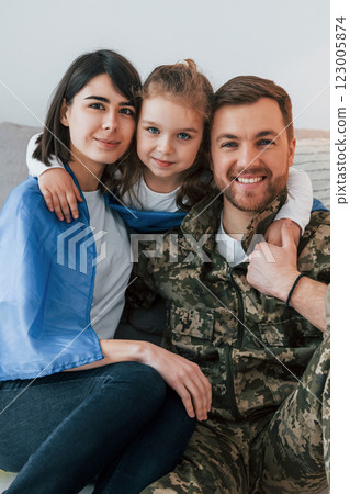 Ukrainian flag is on the shoulders. Soldier in uniform is at home with his wife and daughter 123005874