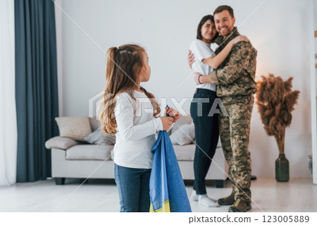 Little girl holding Ukraine flag. Soldier in uniform is at home with his wife and daughter 123005889